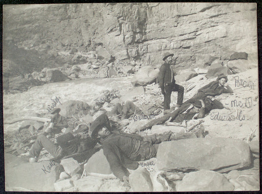 [A Noonday Rest in Marble Canyon, showing well the head of a fall. Snapshot by James Hogue, 1890. From left to right, Travers (standing), Hislop, Kane, Gibson, Ballard, Edwards, Twining (standing), Stanton, McDonald, Brown.]